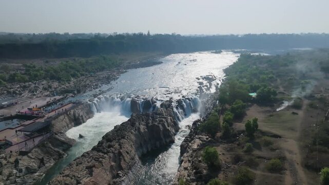 Dhuandhar Falls cascades through rocky gorge with white water rushing over dark basalt cliffs along the Narmada River. Smooth aerial reveals viewing platform, mist rising from falls and Volcanic rock.