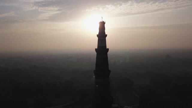 Qutub Minar tower stands silhouetted with camera flare against a hazy sunrise sky in New Delhi, India. Thick smog and air pollution obscure the cityscape surrounding the historic landmark.