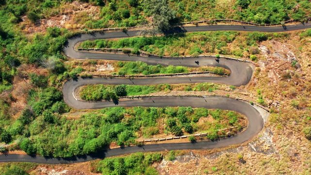 Yercaud's winding mountain road features motorcycles traversing multiple hairpin curves through green and dry terrain. Static aerial view captures the serpentine asphalt road from directly overhead.