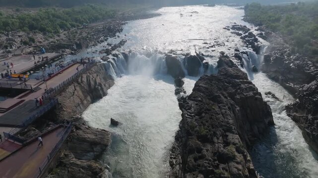 Dhuandhar Falls Narmada river plunges over dark basalt rock formations creating white water rapids through a narrow gorge. Aerial view reveals viewing platforms along the riverbank and volcanic rocks.