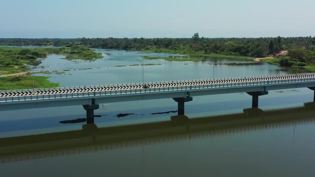 Puducherry's Thirukanji Bridge crosses the calm Gingee river flanked by green wetlands and trees. Smooth tracking shot shows the concrete structure with chevron road markings and a lone motorcycle.