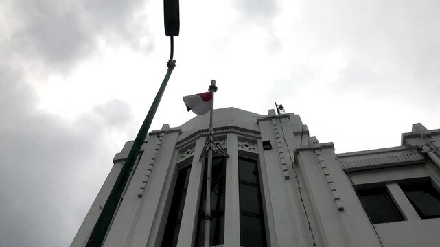 4K handheld low angle of the Indonesian flag waving atop a white colonial building against a cloudy sky. Patriotic historical heritage footage.