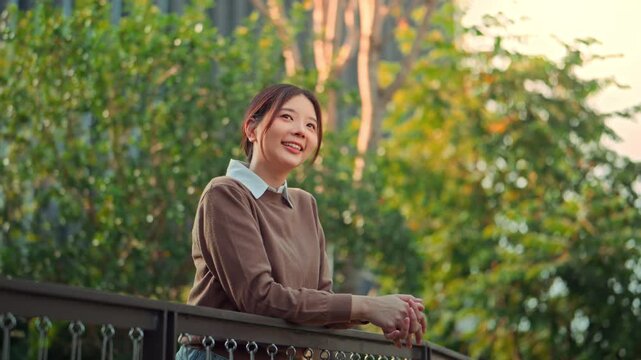 Happy young Asian woman in a brown sweater and white shirt smiling while leaning on a balcony railing in a green park during golden hour. A peaceful female professional enjoying a break outdoors.