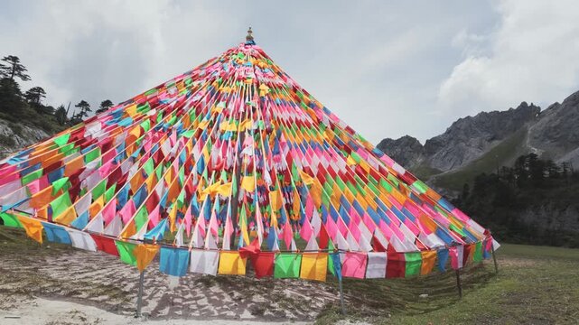 Authentic Tibetan prayer flags arranged in a pyramid shape wave in the wind against a backdrop of rugged mountains. Useful for travel documentaries and cultural wellness content.