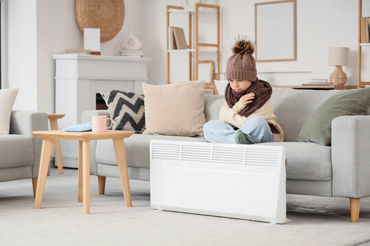 Frozen little girl sitting on sofa near electric convector heater at home