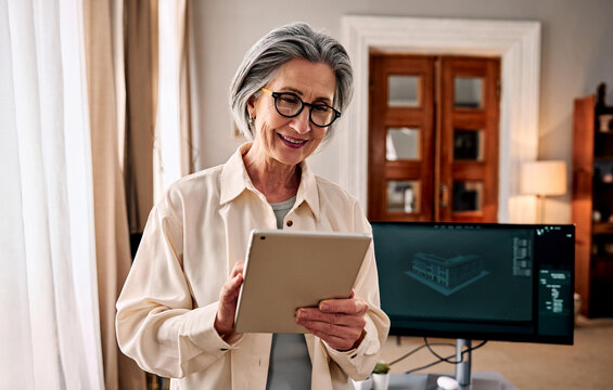 Senior woman using tablet in home office with computer monitor.