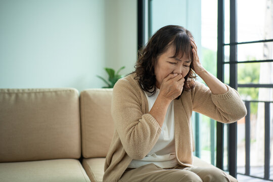 Unhappy Asian senior woman coughing with illness and fatigue while sitting alone on living room sofa, healthcare and medical protection planning for life insurance in retirement age