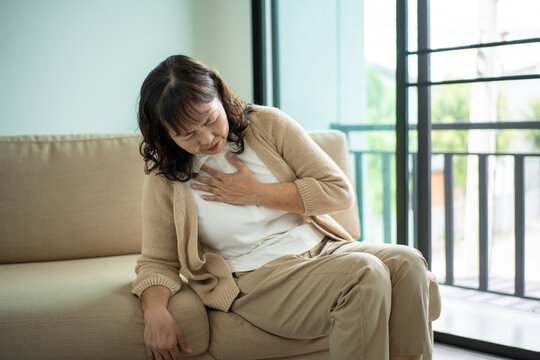 Unhappy Asian senior woman coughing with illness and fatigue while sitting alone on living room sofa, healthcare and medical protection planning for life insurance in retirement age