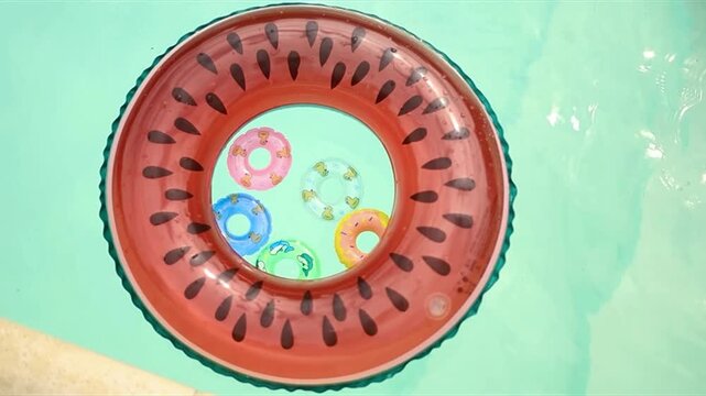 Watermelon and small donut inflatable rings gently floating in a clear blue swimming pool, top-down