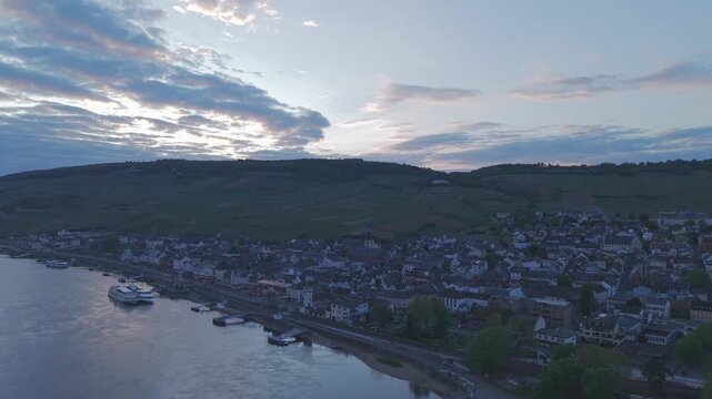 Beautiful aerial drone footage of Rudesheim am Rhein, Germany during sunset and blue hour. Cinematic view of the Rhine River waterfront, traditional German town, vineyard hills and reflective water un