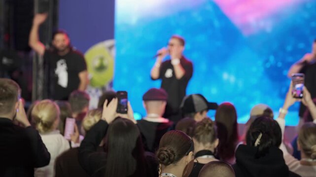 Crowd of fans recording live performance with smartphones, blurred stage lights and energetic atmosphere.