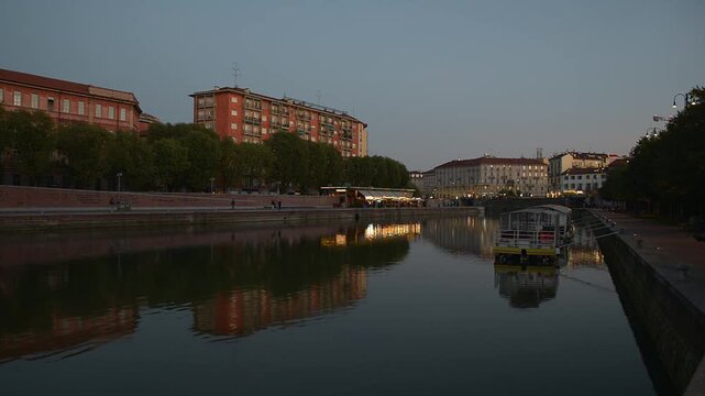 Milan Navigli Darsena at dusk, waterfront canal in Porta Ticinese area with calm water reflections, cityscape lights, small boat, people walking promenade, Milano, Italy