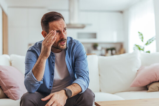 Stressed Man Sitting On Sofa Holding Head In Modern Living Room With Worried Expression