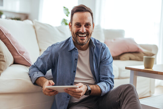 Smiling Man Using Tablet At Home In Bright Living Room, Casual Remote Work And Relaxation