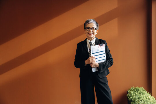 Senior businesswoman in formal black suit holding documents and smiling, standing against a brown wall with a small green plant in the foreground