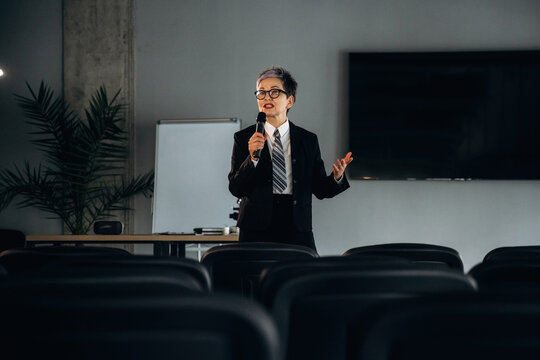 Business professional in black suit delivers presentation with microphone in modern office setting, chairs arranged in rows and large screen visible in background