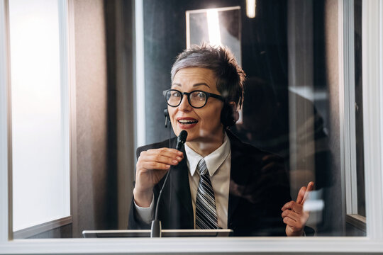 Female interpreter wearing headset speaks into microphone inside soundproof booth, with glass walls and professional office environment visible in the background