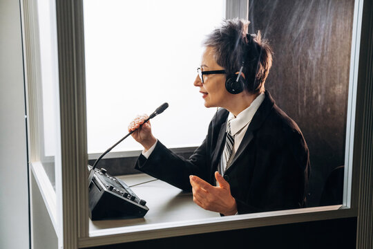 Female interpreter wearing headphones speaks into a microphone inside a soundproof booth with a control panel and minimalistic interior design visible