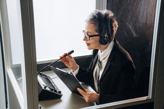 Female interpreter wearing headset speaks into microphone while seated in soundproof booth, holding clipboard and surrounded by audio equipment in a modern office setting