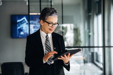 Fototapeta premium Businesswoman in formal attire using a tablet while standing in a modern office with glass walls and a digital presentation displayed in the background