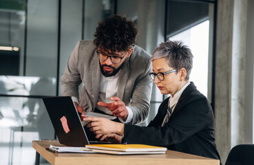 Obraz premium Business professionals collaborating on a laptop in a modern office setting, with documents and a wooden table visible in the foreground and large windows in the background