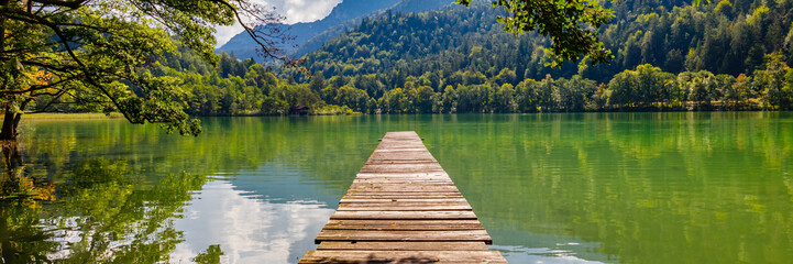 Naklejka premium Wooden pier extends into green lake surrounded by mountains and trees during sunny day
