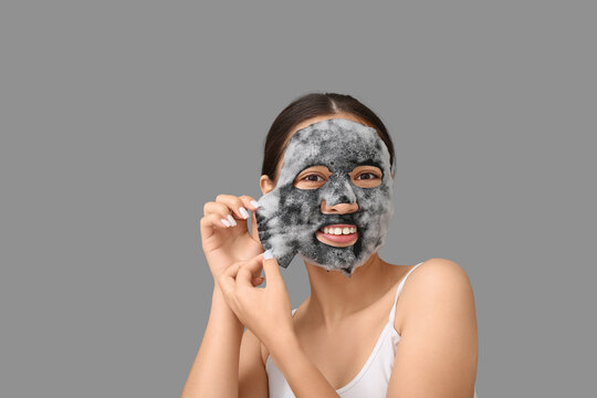Young woman removing activated charcoal mask from her face against grey background