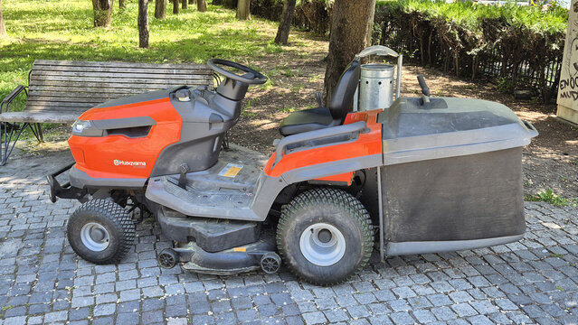 An orange and grey Husqvarna riding lawn mower with a large rear grass collector parked on a cobblestone path in a park. Picture taken on May 7, 2026 in Bucharest, Romania.