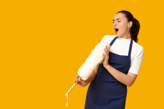 Portrait of beautiful young housewife in apron using pp duster as microphone on yellow background