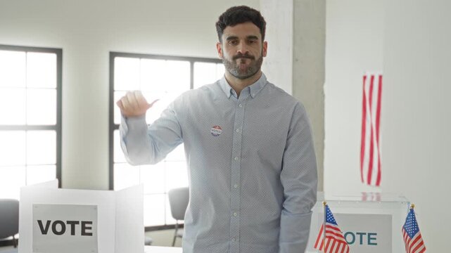 Man wearing i voted sticker gives thumbs down beside a clear ballot box and voting booth in a polling area inside a building; frustration.