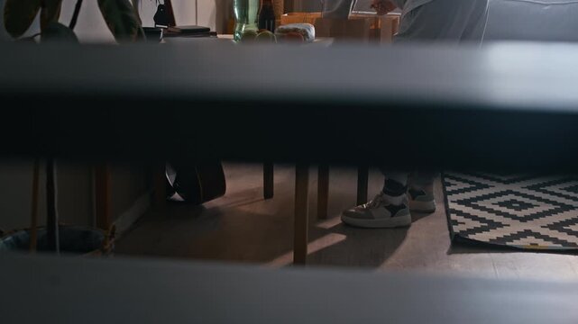 Tilt up of Biracial woman wearing electronic ankle monitor unpacking grocery bags and arranging household products on table in home interior