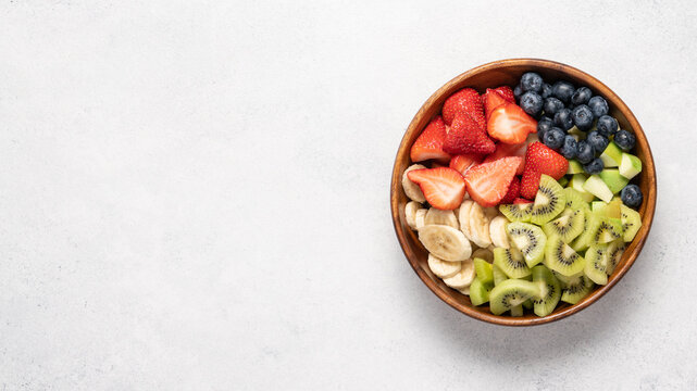 Fresh fruit salad with banana apple kiwi blueberry and strawberry in wooden bowl, top view on light background, healthy eating concept