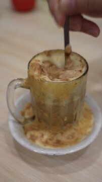 Close-up vertical footage of traditional Indonesian Teh Talua being stirred in a glass cup at a cozy cafe. Creamy Minangkabau egg tea foam with warm texture and shallow depth of field.
