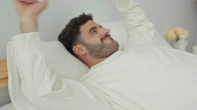 Man face smiling, stretches arms overhead on white sheets and pillow in bright bedroom with wooden headboard and vase of yellow flowers; morning calm contentment.