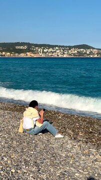 Woman on pebble beach with phone in hand by turquoise sea in Nice, French Riviera.