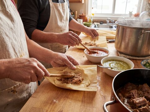 Mexican hands assembling traditional tamales corn husks masa dough close cooking authentic preparation - skilled adult wrapping flavorful filling using ancestral cultural technique on rustic wooden