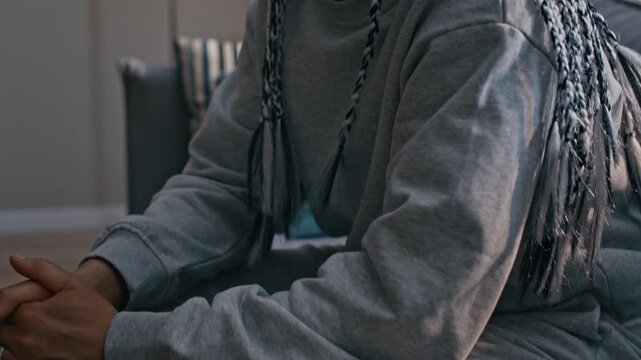 Tilt up shot of Biracial woman in grey sports suit sitting on couch with electronic monitoring bracelet on her ankle, looking stressed and wiping her face in frustration
