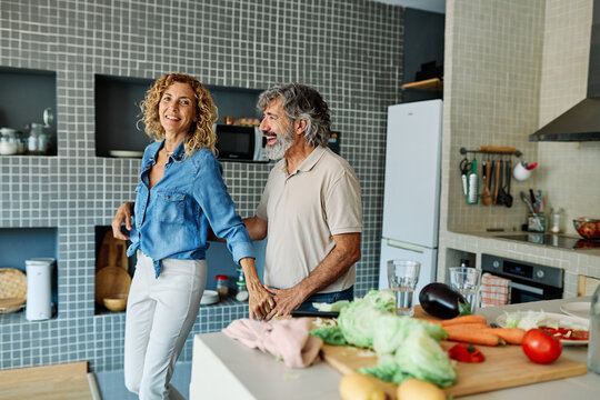 Portrait of happy senior mid aged mature couple prepering meal and having fun dancing and embracing in kitchen at hpme