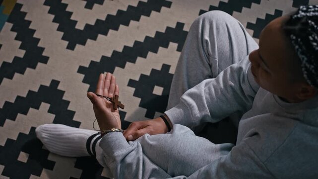 From above shot of Biracial woman sitting on apartment floor holding cross in prayer, seeking comfort and hope during difficult home confinement situation