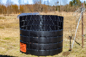 Obraz premium Black circular compost bin with orange door, made of plastic panels, standing outdoors on grass, ready for organic waste processing.