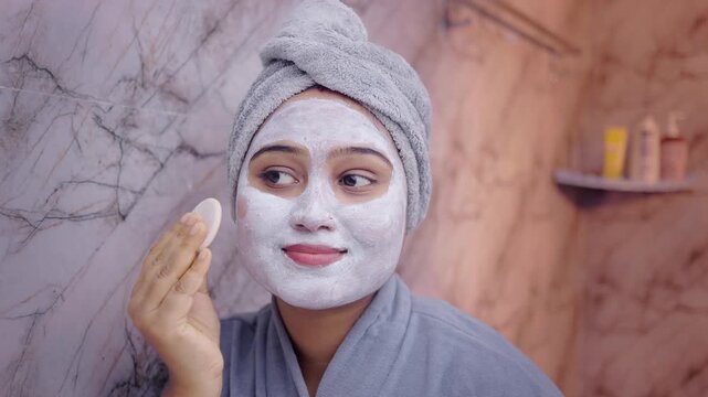 Close up shot of a young woman gently removing face pack with a cotton pad while looking into the bathroom mirror, showcasing daily skincare routine and self-care.