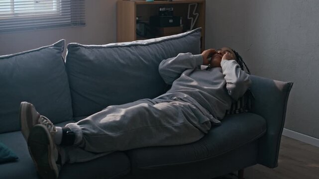 Eye level shot of Biracial female wearing grey outfit and ankle bracelet lying on sofa at home, appearing frustrated and emotionally overwhelmed