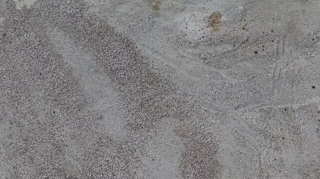 Drone top-down view over sand and gravel surface in Essendon, Hertfordshire, England. Forward motion reveals abstract ground texture, construction aggregate patterns, and subtle tire marks.