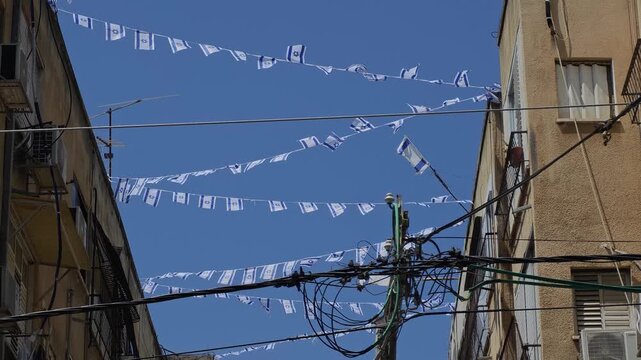 Upward street view of aged buildings connected by strings of Israeli flags fluttering in wind, worn facades framing bright blue sky, urban heritage atmosphere, patriotic scene, horizontal composition