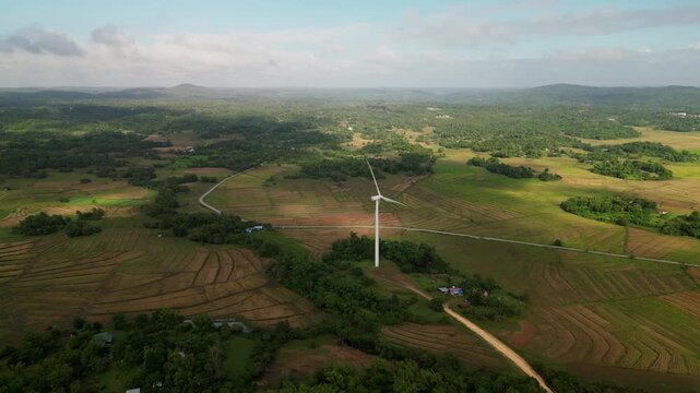 Aerial flyover of a single wind turbine rising above patchwork farmland and rolling hills in Guimaras, Visayas, Philippines.