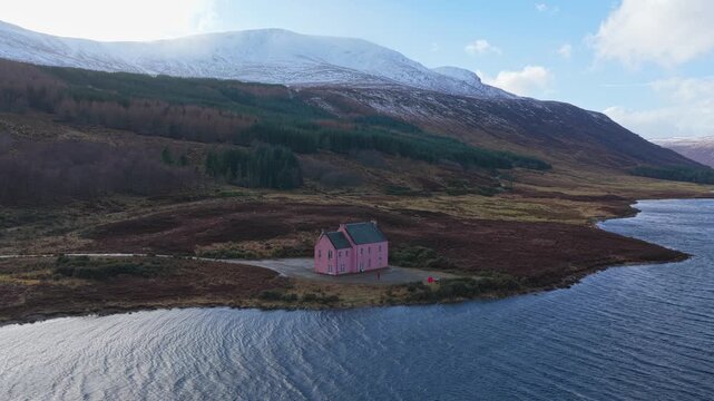 Aerial view of a distinctive pink house, known as a bothy, situated by a tranquil loch with snowy mountains in the background