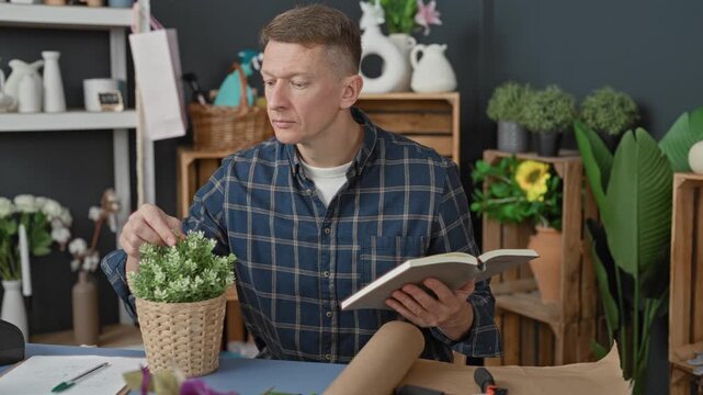 Young man adjusts potted plant, hand touching leaves while holding open book and checking notes on table in a florist studio; careful concentration.