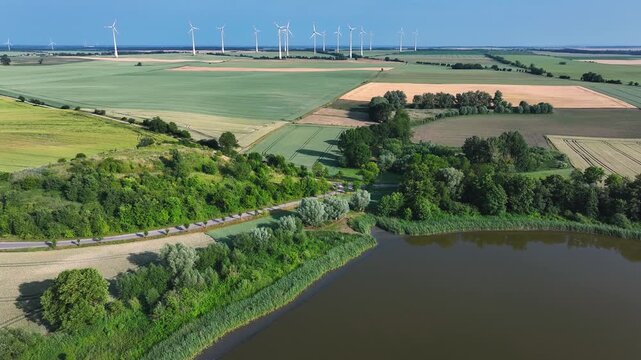 Aerial view of wind turbines standing over vast agricultural fields and a calm lake under a clear blue sky in Uckermark, Brandenburg, Germany.