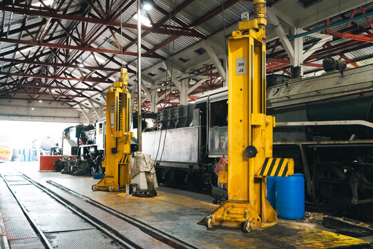 Engineers vehicle transportation logistic train maintenance workshop interior with vintage locomotive under repair and tall yellow jack lifting system creating busy industrial atmosphere