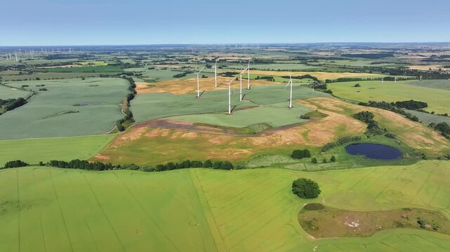 Aerial view of wind turbines standing in a vast rural landscape with green fields and a small pond under a clear blue sky, Uckermark, Brandenburg, Germany.